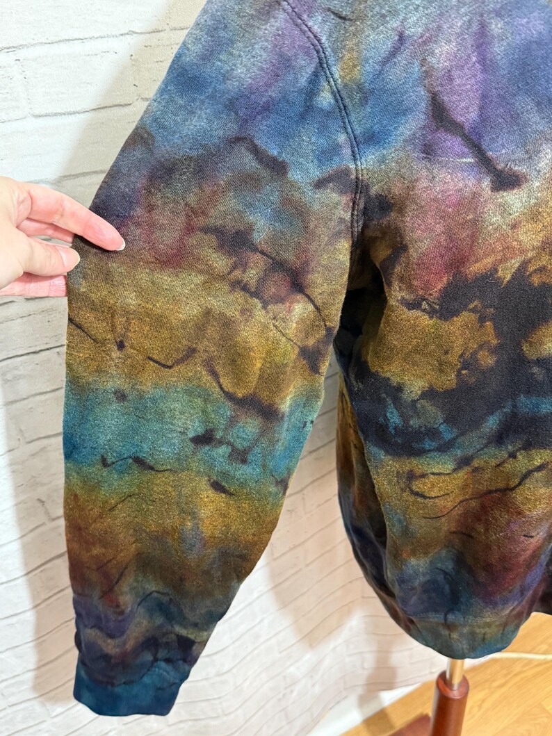 May include: A tie-dye sweatshirt in shades of blue, purple, gold, and brown. The garment has a unique, marbled effect, with the colors blending and swirling together. The sleeve is visible, showcasing the intricate dye pattern.