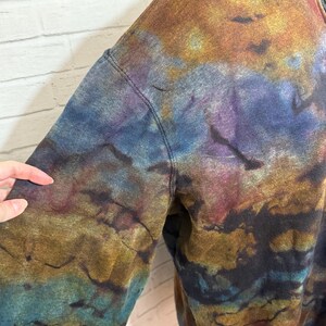 May include: A tie-dye sweatshirt in shades of blue, gold, purple, and brown. The garment has a zipper and a stitched seam. The fabric has a textured appearance, with the colors blending together in a marbled effect.