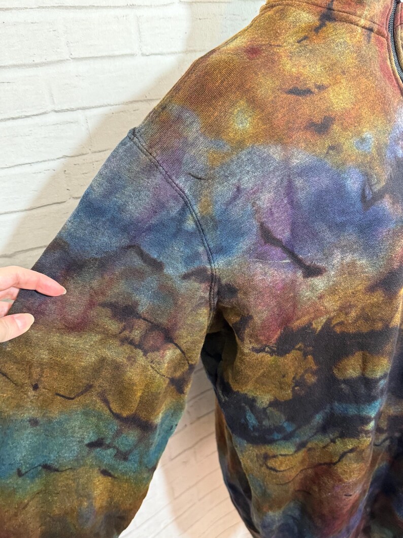 May include: A tie-dye sweatshirt in shades of blue, gold, purple, and brown. The garment has a zipper and a stitched seam. The fabric has a textured appearance, with the colors blending together in a marbled effect.