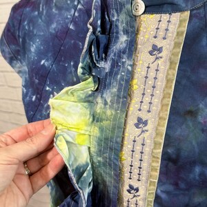May include: A tie-dye short-sleeve top in shades of blue, purple, and yellow. The top features a decorative floral ribbon and a single button closure. The ruffled trim adds a unique design element to the garment.