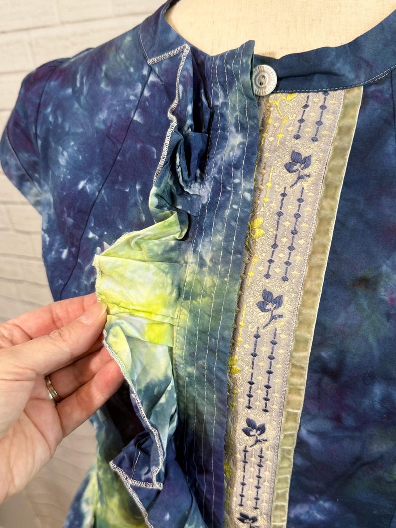 May include: A tie-dye short-sleeve top in shades of blue, purple, and yellow. The top features a decorative floral ribbon and a single button closure. The ruffled trim adds a unique design element to the garment.