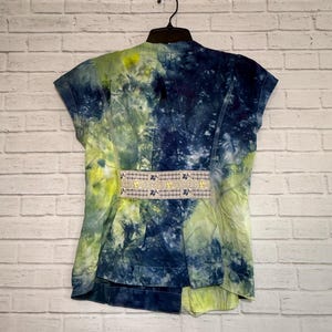 May include: A short-sleeved jacket with a tie-dye pattern in shades of blue and yellow-green. The jacket has a decorative band with a floral design at the waist. The garment is hanging on a black hanger against a white brick wall.