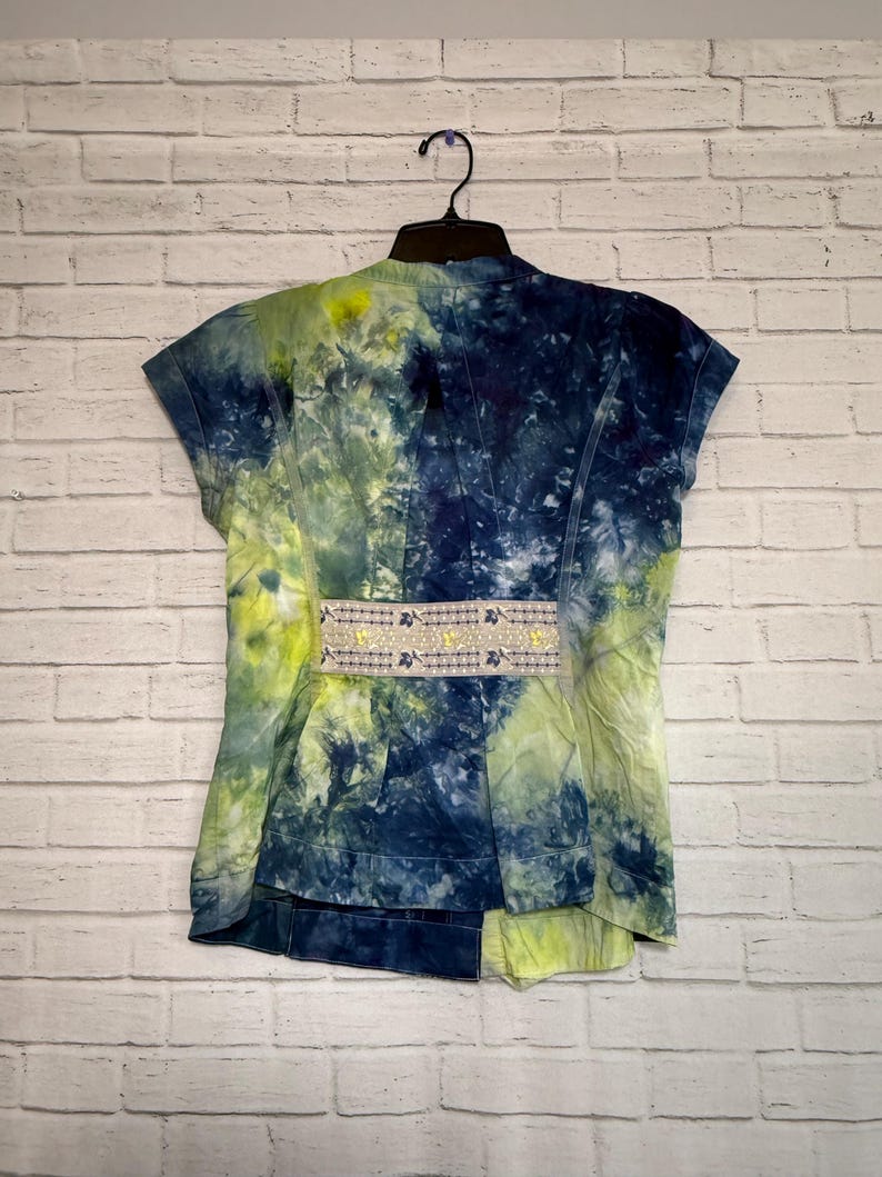May include: A short-sleeved jacket with a tie-dye pattern in shades of blue and yellow-green. The jacket has a decorative band with a floral design at the waist. The garment is hanging on a black hanger against a white brick wall.