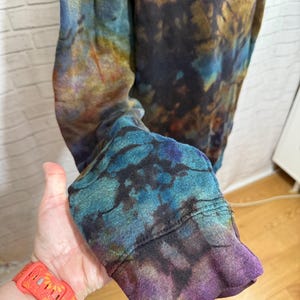 May include: A long-sleeved shirt with a tie-dye pattern in shades of blue, purple, and gold. The cuff of the sleeve is visible, showcasing the vibrant colors and unique design. The shirt appears to be made of a soft, comfortable material.