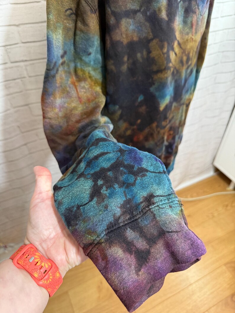 May include: A long-sleeved shirt with a tie-dye pattern in shades of blue, purple, and gold. The cuff of the sleeve is visible, showcasing the vibrant colors and unique design. The shirt appears to be made of a soft, comfortable material.