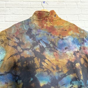 May include: A tie-dye fleece pullover with a high collar. The fabric features a blend of colors including blue, brown, orange, and black. The garment is hanging on a wooden hanger against a white brick wall.