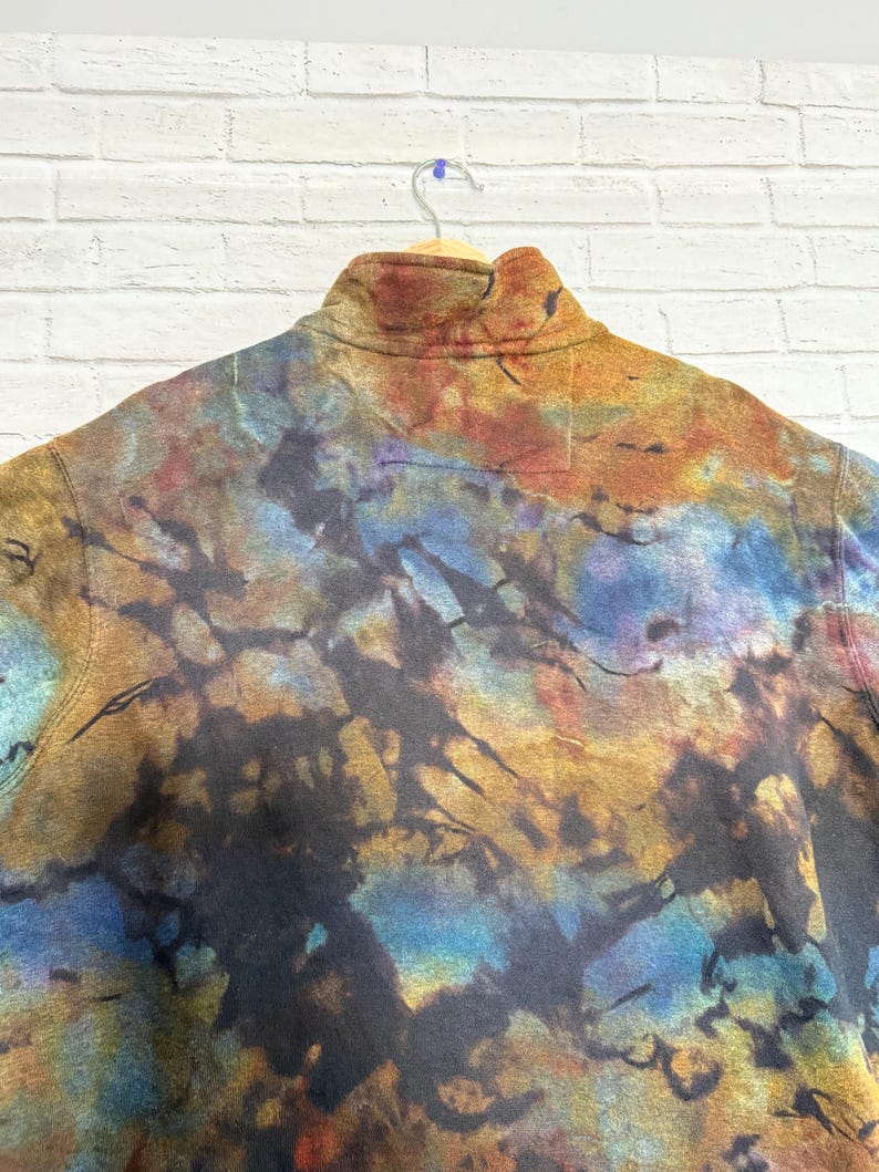 May include: A tie-dye fleece pullover with a high collar. The fabric features a blend of colors including blue, brown, orange, and black. The garment is hanging on a wooden hanger against a white brick wall.