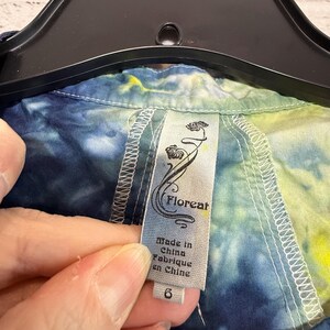 May include: A close-up of a tie-dye shirt with a tag that reads "Floreat" and "Made in China". The shirt is a mix of blue, yellow, and green colors. The tag also indicates a size 6.