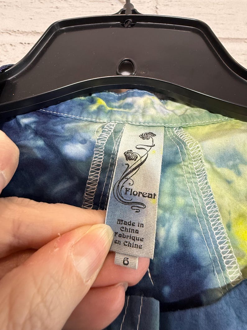 May include: A close-up of a tie-dye shirt with a tag that reads "Floreat" and "Made in China". The shirt is a mix of blue, yellow, and green colors. The tag also indicates a size 6.
