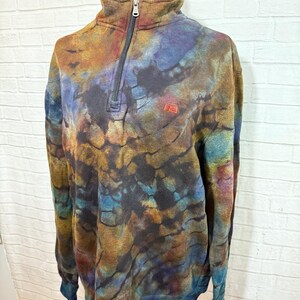 May include: A tie-dye pullover sweatshirt with a quarter-zip neckline. The colors include shades of brown, blue, and orange. The sweatshirt has a small red logo on the chest and a ribbed collar and cuffs.