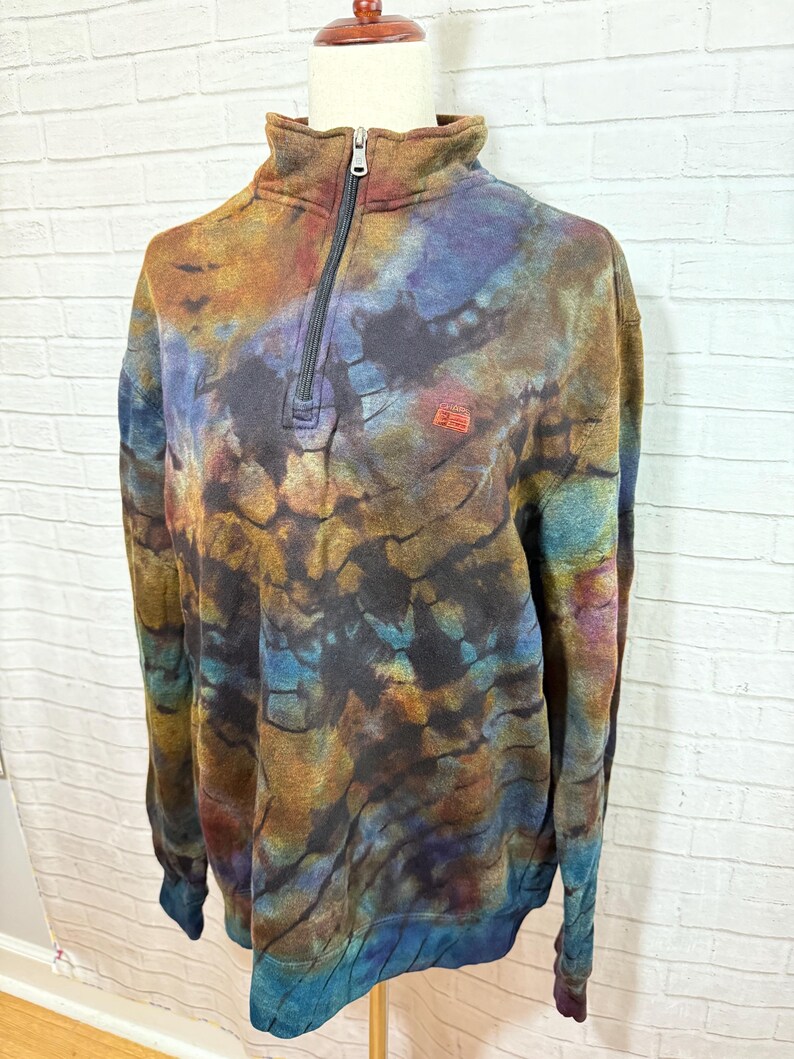 May include: A tie-dye pullover sweatshirt with a quarter-zip neckline. The colors include shades of brown, blue, and orange. The sweatshirt has a small red logo on the chest and a ribbed collar and cuffs.