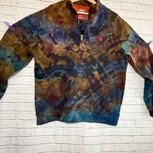May include: A tie-dye Chaps sweatshirt with a quarter-zip collar. The sweatshirt features a blend of brown, blue, and purple hues. The Chaps logo is visible on the chest. The sweatshirt is hanging on a wooden hanger.