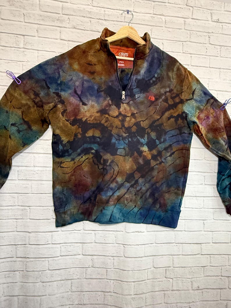 May include: A tie-dye Chaps sweatshirt with a quarter-zip collar. The sweatshirt features a blend of brown, blue, and purple hues. The Chaps logo is visible on the chest. The sweatshirt is hanging on a wooden hanger.