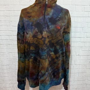 May include: A tie-dye quarter-zip sweatshirt in shades of brown, blue, and purple. The sweatshirt has a high collar and a small red logo on the chest. The zipper is black.