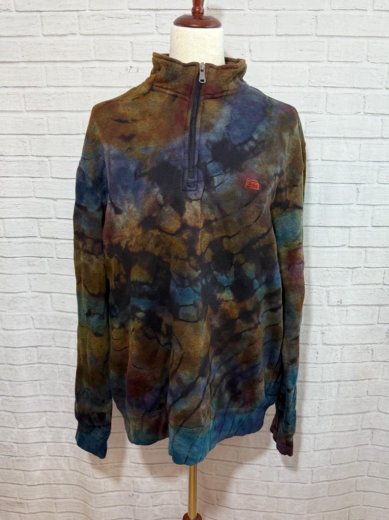 May include: A tie-dye quarter-zip sweatshirt in shades of brown, blue, and purple. The sweatshirt has a high collar and a small red logo on the chest. The zipper is black.