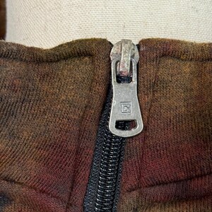 May include: Close-up of a brown and red tie-dye zip-up hoodie. The silver zipper is centered, with a black zipper track. The fabric has a textured appearance. The hoodie is displayed on a mannequin.