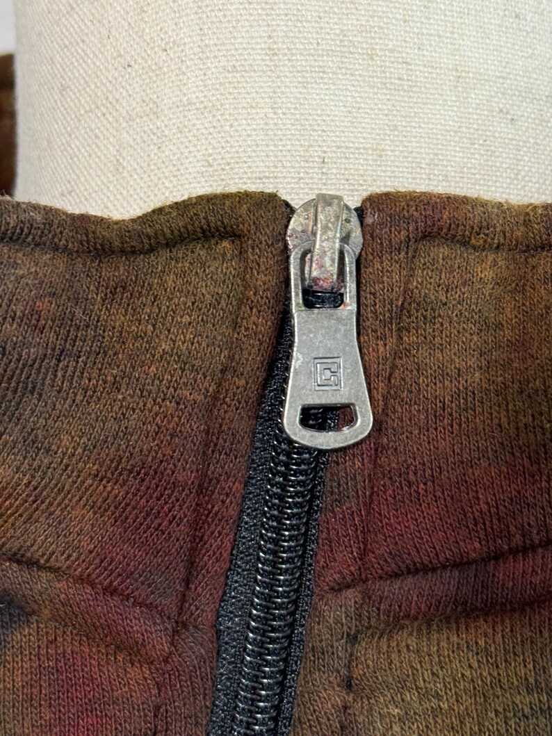 May include: Close-up of a brown and red tie-dye zip-up hoodie. The silver zipper is centered, with a black zipper track. The fabric has a textured appearance. The hoodie is displayed on a mannequin.