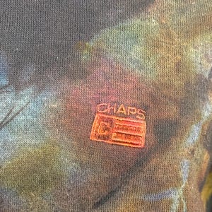 May include: Close-up of a dark blue, tie-dye patterned sweatshirt. The fabric has a textured appearance with a colorful blend of blues, greens, and pinks. A small, embroidered "CHAPS" logo in red is visible.