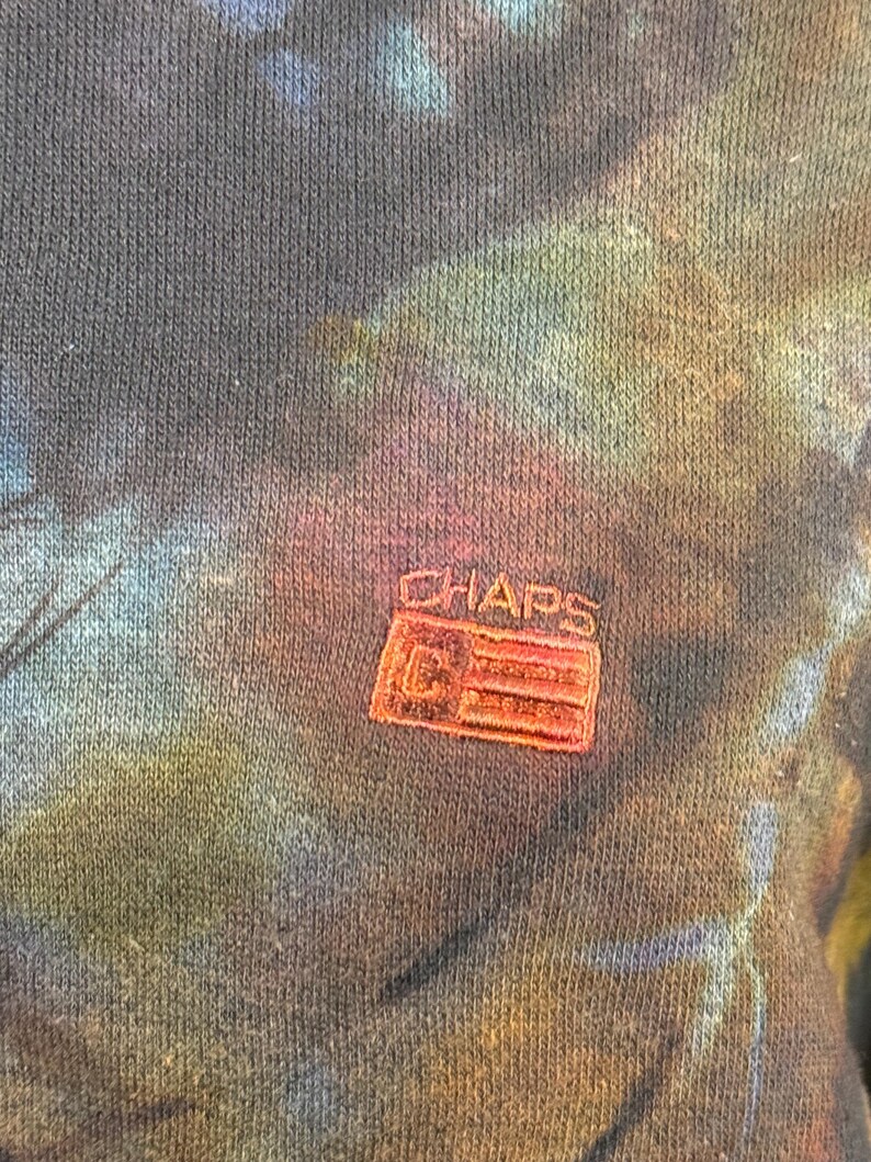 May include: Close-up of a dark blue, tie-dye patterned sweatshirt. The fabric has a textured appearance with a colorful blend of blues, greens, and pinks. A small, embroidered "CHAPS" logo in red is visible.