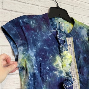 May include: A short-sleeved tie-dye shirt in shades of blue, purple, and yellow. The shirt has a ruffled trim down the front and a decorative embroidered panel. The shirt is hanging on a black hanger against a white brick wall.