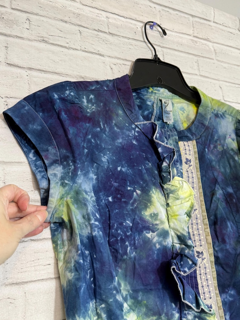 May include: A short-sleeved tie-dye shirt in shades of blue, purple, and yellow. The shirt has a ruffled trim down the front and a decorative embroidered panel. The shirt is hanging on a black hanger against a white brick wall.