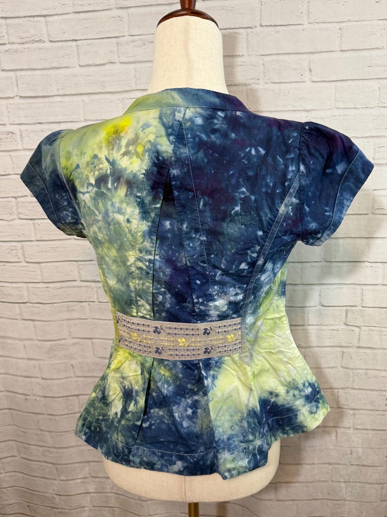 May include: A short-sleeved jacket with a tie-dye pattern in shades of blue, yellow, and purple. The jacket has a fitted waist with a decorative belt and a peplum hem. The fabric appears to be cotton or a similar material.