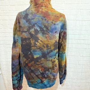 May include: A tie-dye fleece pullover with a high collar, displayed on a mannequin. The colors include shades of blue, brown, gold, and purple, creating a unique, abstract pattern. The long-sleeved top has a relaxed fit.