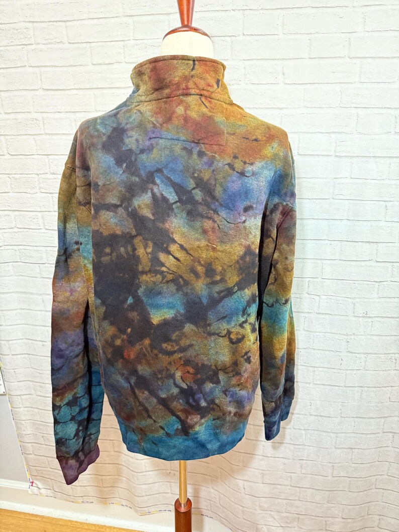 May include: A tie-dye fleece pullover with a high collar, displayed on a mannequin. The colors include shades of blue, brown, gold, and purple, creating a unique, abstract pattern. The long-sleeved top has a relaxed fit.