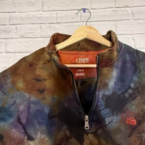 May include: A tie-dye patterned fleece pullover with a quarter-zip closure. The top features the "CHAPS" logo on the chest and a tag at the neckline. The colors include brown, blue, and orange.