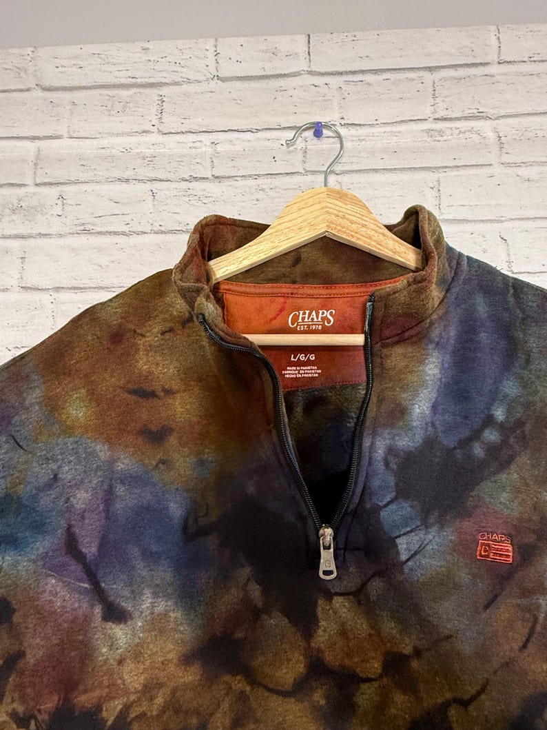 May include: A tie-dye patterned fleece pullover with a quarter-zip closure. The top features the "CHAPS" logo on the chest and a tag at the neckline. The colors include brown, blue, and orange.