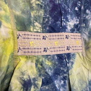 May include: Close-up of a tie-dye fabric in shades of blue, green, and yellow. A decorative band with floral and leaf patterns in yellow and blue accents the garment. The fabric appears to be a dress or top.