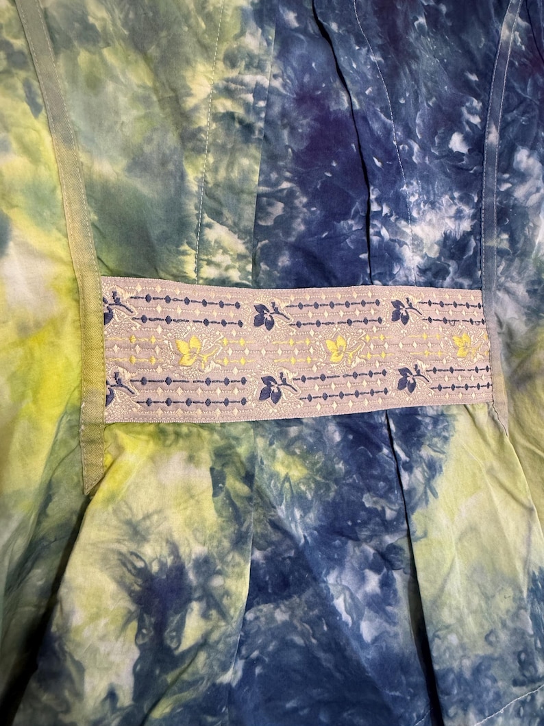 May include: Close-up of a tie-dye fabric in shades of blue, green, and yellow. A decorative band with floral and leaf patterns in yellow and blue accents the garment. The fabric appears to be a dress or top.