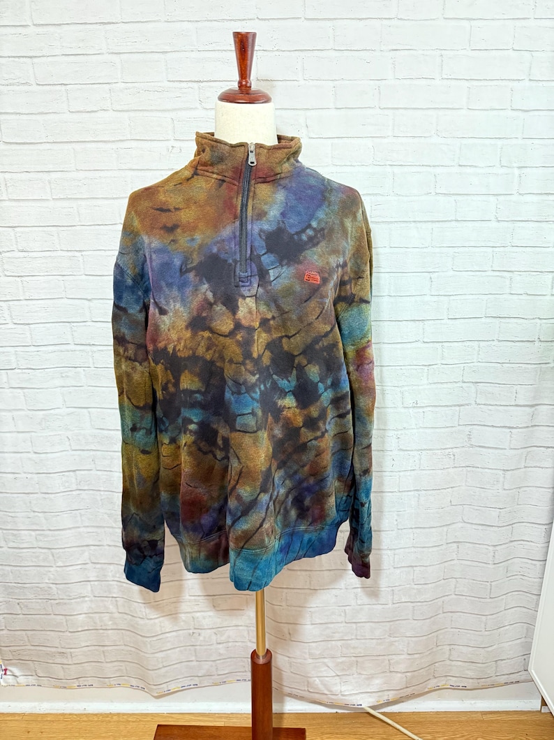 May include: A tie-dye pullover sweatshirt with a quarter-zip closure and a small red logo. The sweatshirt features a mix of colors, including brown, blue, and teal. The garment is displayed on a mannequin against a white brick wall.
