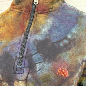 May include: A tie-dye style sweatshirt with a quarter-zip closure. The sweatshirt features a mix of colors including brown, blue, and purple. The brand logo "CHAPS" is visible on the lower right side of the sweatshirt.