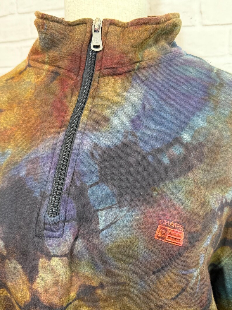 May include: A tie-dye style sweatshirt with a quarter-zip closure. The sweatshirt features a mix of colors including brown, blue, and purple. The brand logo "CHAPS" is visible on the lower right side of the sweatshirt.