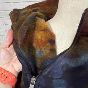 May include: A tie-dye hoodie with a half-zip closure. The colors include shades of brown, orange, blue, and purple. The interior of the hood is a lighter shade of orange. The zipper is black with a silver pull.