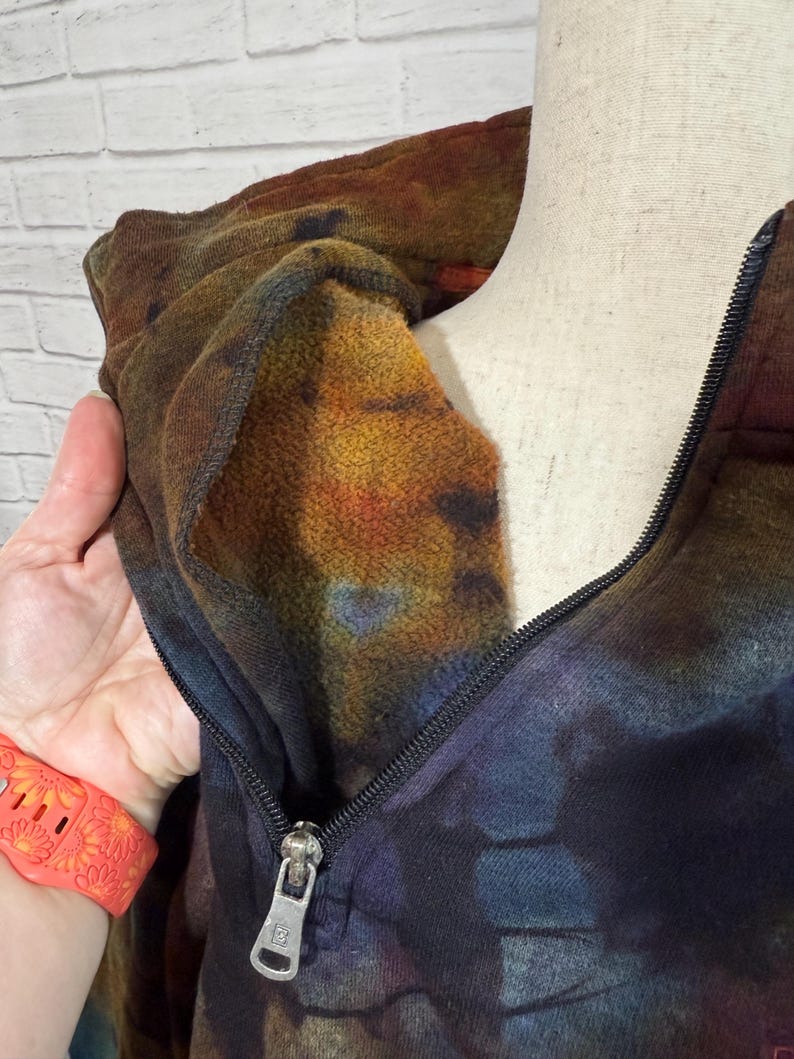 May include: A tie-dye hoodie with a half-zip closure. The colors include shades of brown, orange, blue, and purple. The interior of the hood is a lighter shade of orange. The zipper is black with a silver pull.