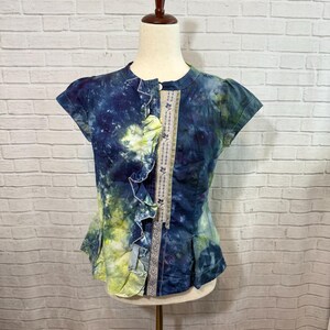 May include: A short-sleeved, tie-dye top in shades of blue and yellow. The top features a ruffled front and a decorative panel with floral embroidery. The garment is displayed on a mannequin, showcasing its unique design and style.