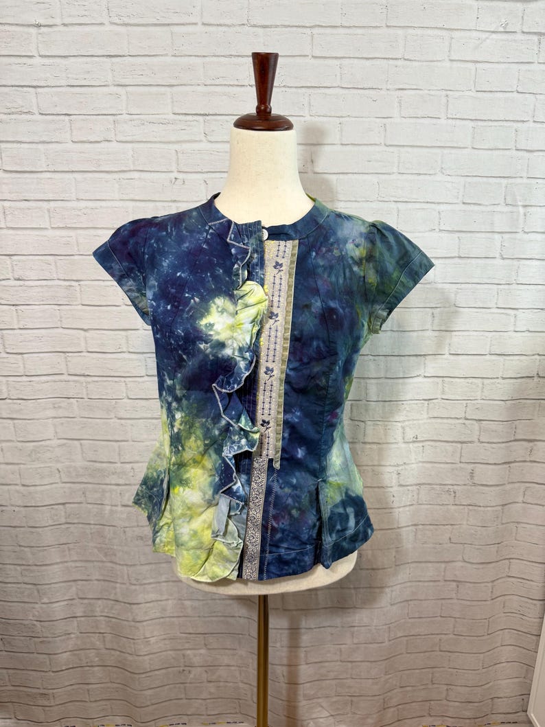 May include: A short-sleeved, tie-dye top in shades of blue and yellow. The top features a ruffled front and a decorative panel with floral embroidery. The garment is displayed on a mannequin, showcasing its unique design and style.