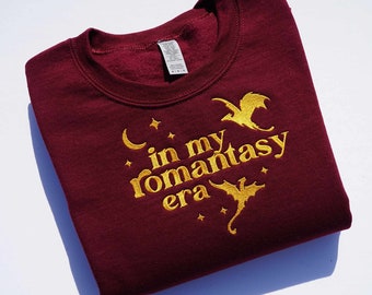 May include: Burgundy sweatshirt with gold embroidery that reads "in my romantasy era" with a crescent moon and stars. Two gold dragons are embroidered on either side of the text.