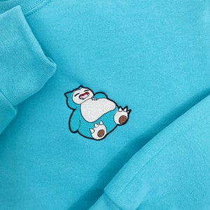 Pu&ograve; includere: Una felpa di colore azzurro con un disegno ricamato di un Snorlax blu e marrone che dorme sul davanti.