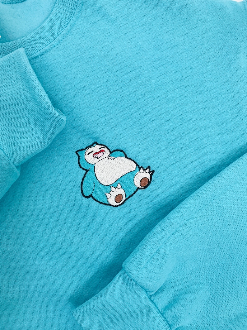 Pu&ograve; includere: Una felpa di colore azzurro con un disegno ricamato di un Snorlax blu e marrone che dorme sul davanti.
