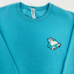Pu&ograve; includere: Una felpa di colore blu turchese con un'immagine ricamata di un personaggio Pokemon blu e bianco in stile cartone animato sul petto sinistro.