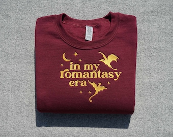 May include: Burgundy sweatshirt with gold embroidery that reads "in my romantasy era" with two dragons and stars.