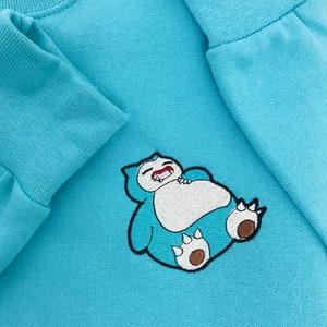 Pu&ograve; includere: Una felpa di colore azzurro chiaro con un'immagine ricamata di un personaggio dei cartoni animati blu e bianco con la bocca spalancata. Il personaggio &egrave; sdraiato sulla schiena con le braccia e le gambe distese.