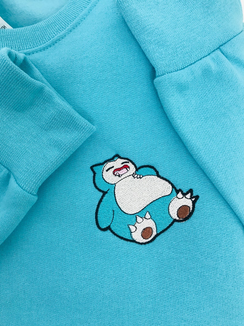Pu&ograve; includere: Una felpa di colore azzurro chiaro con un'immagine ricamata di un personaggio dei cartoni animati blu e bianco con la bocca spalancata. Il personaggio &egrave; sdraiato sulla schiena con le braccia e le gambe distese.