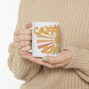 Op de afbeelding: Een witte keramische mok met een retro zonnestralenontwerp. De mok heeft de tekst "Caffeine & Cream" in geel.