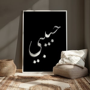 May include: A framed art print featuring the Arabic word "Habibi" in a 3D silver metallic font against a black background. The artwork is displayed in a room with a neutral color palette, a woven basket, and a patterned rug.
