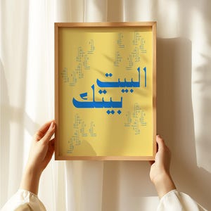 Könnte beinhalten: Ein rechteckiger Wandkunstdruck in einem hellen Holzrahmen mit gelbem Hintergrund und blauer arabischer Kalligrafie. Der zentrale Text lautet Al-Bayt Baytak, umgeben von kleineren, sich wiederholenden blauen arabischen Textmustern.