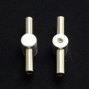 May include: Two brushed metal cabinet pulls. Each pull has a cylindrical handle extending from a circular centre piece. One pull has a flat centre, while the other has a recessed screw. The background is black.